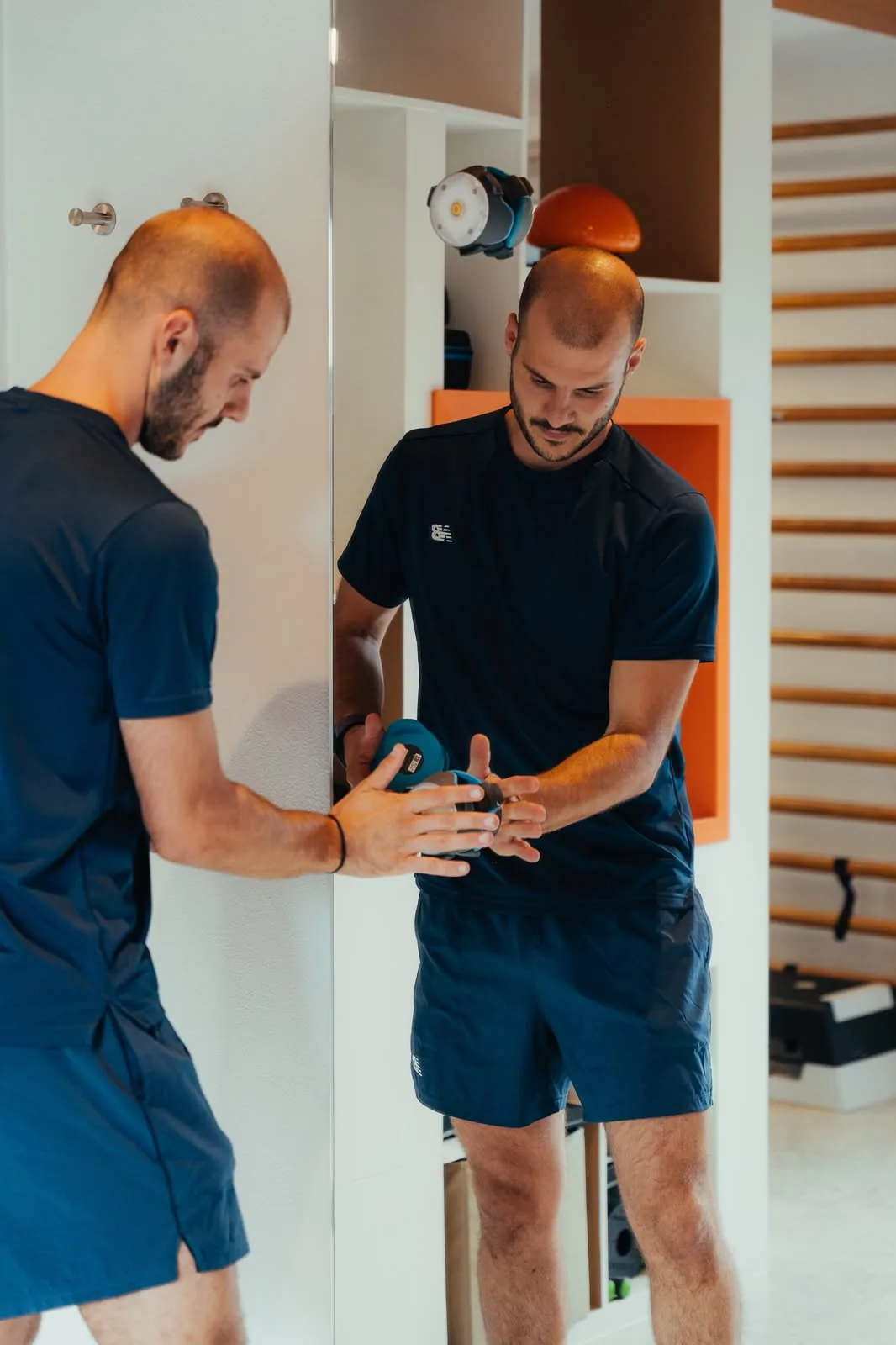 Deux hommes en tenue de sport face à face, l'un tient une balise lumineuse, devant un espalier.